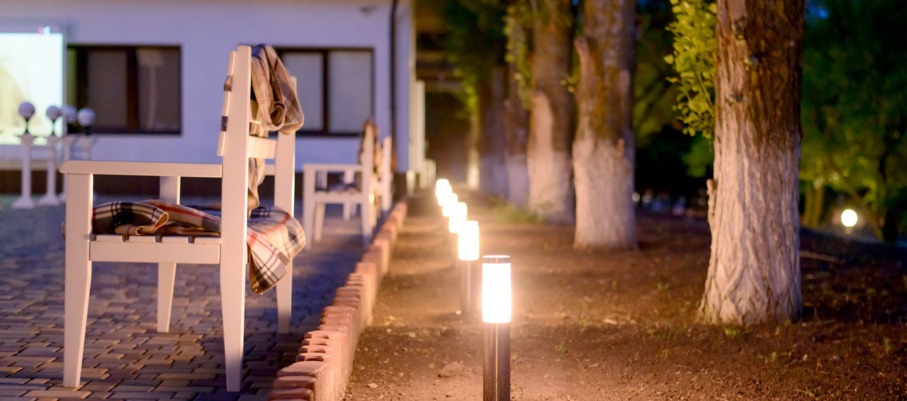 White chairs draped with blankets outside a modern home with a row of trees and solar lighting. 