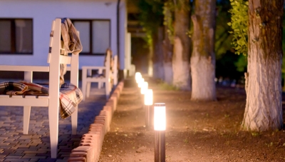 White chairs draped with blankets outside a modern home with a row of trees and solar lighting. 